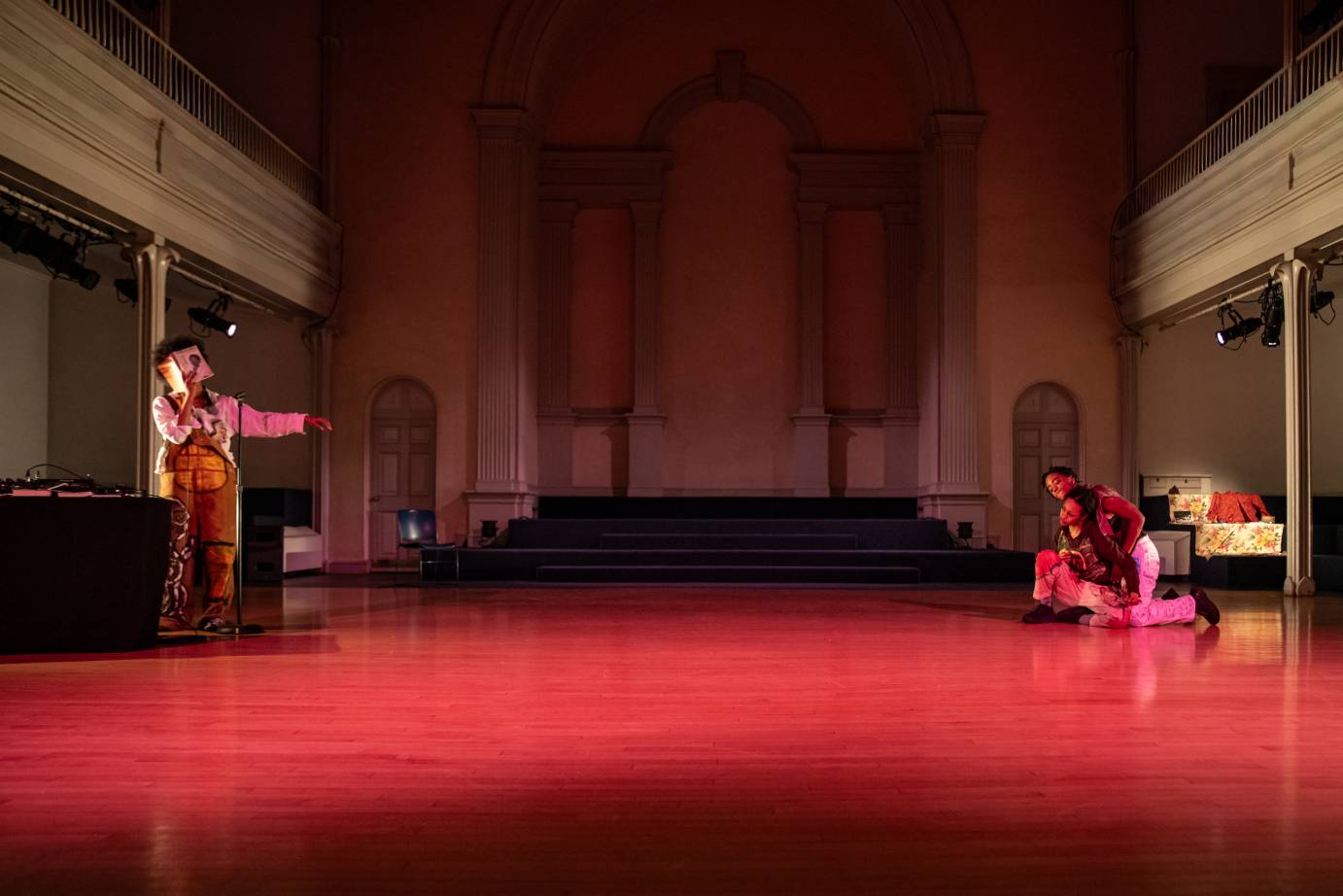 One performer reads from a book while the other two are intwined on the other side of the stage. They are bathed in red light.